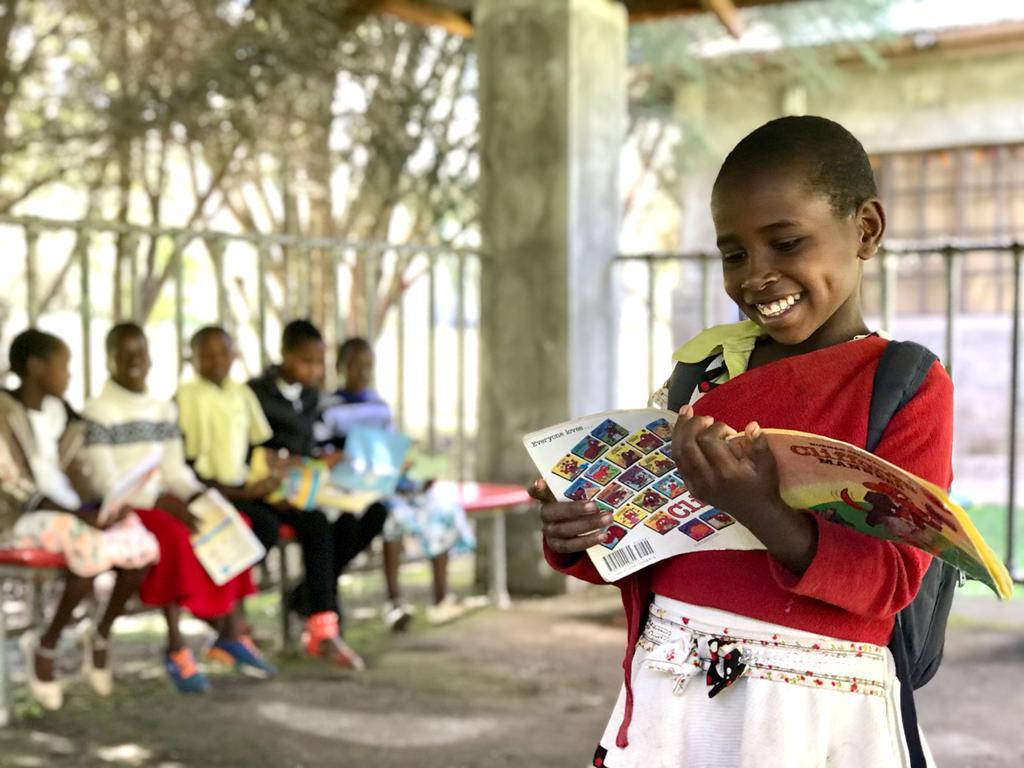 A smiling child reading a book while other children sit together in the background.
